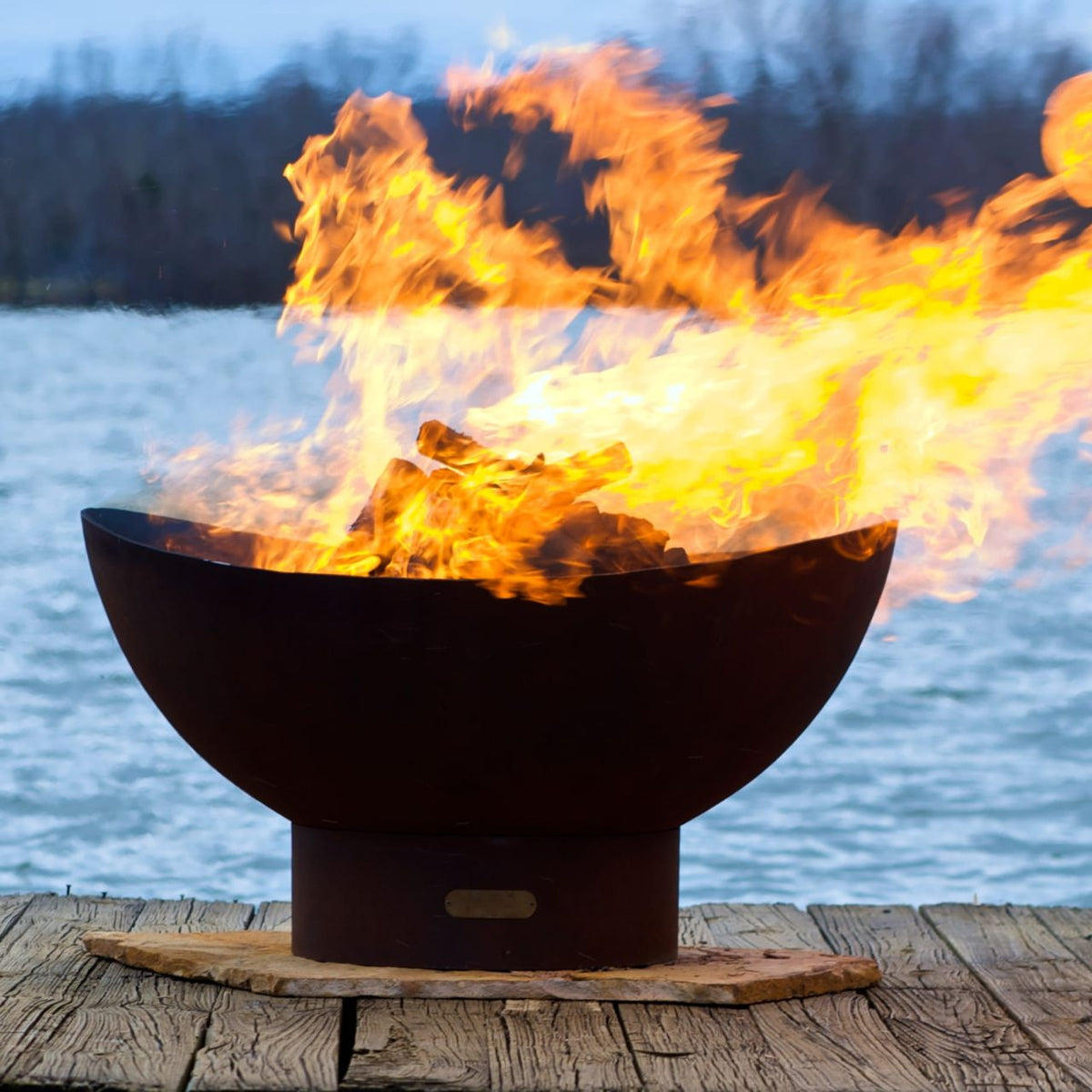 Scallop 36-inch carbon steel fire pit roaring with flames on a dock, set against a peaceful lake with trees in the background. Made in USA by Fire Pit Art.