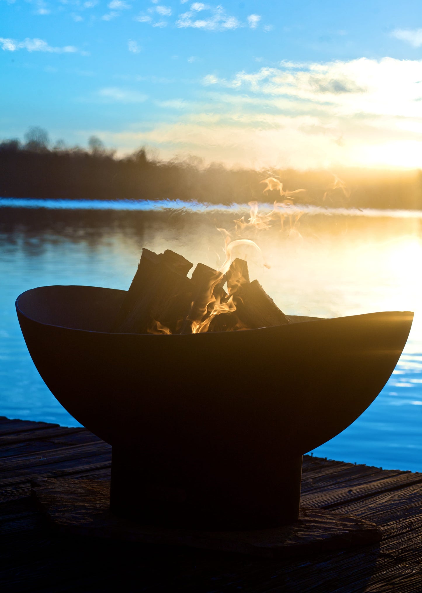 Scallop fire pit glowing at sunset on a dock, with flames flickering in silhouette against a reflective lake and golden sky.