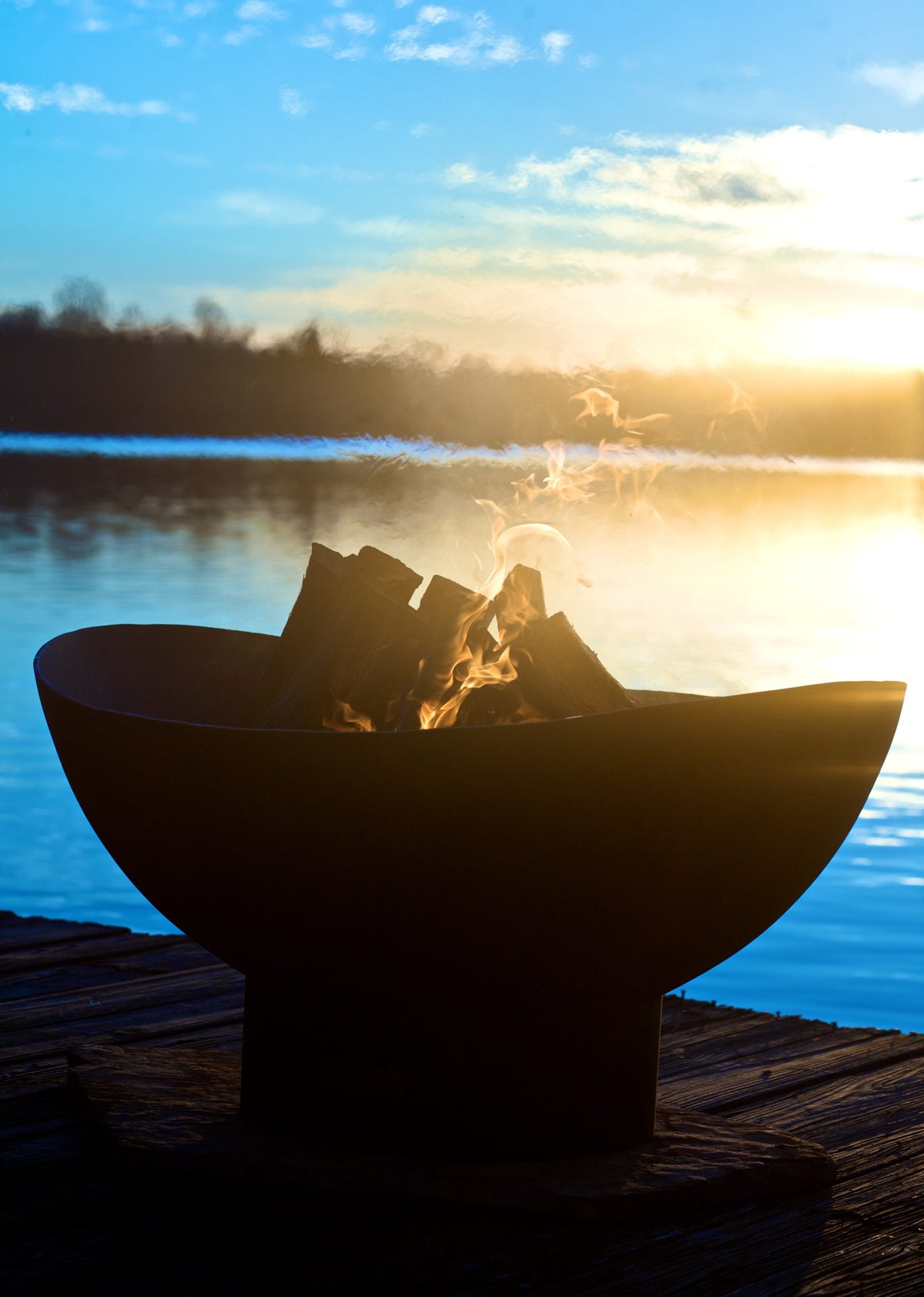 Scallop fire pit glowing at sunset on a dock, with flames flickering in silhouette against a reflective lake and golden sky.