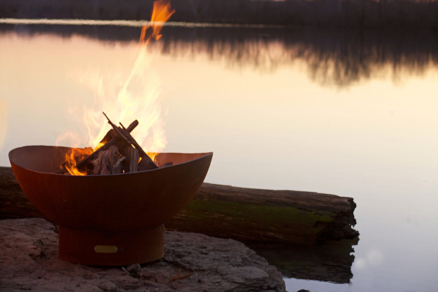 Scallop 36" carbon steel fire pit burning on a stone surface by the water with a reflection and driftwood log nearby.