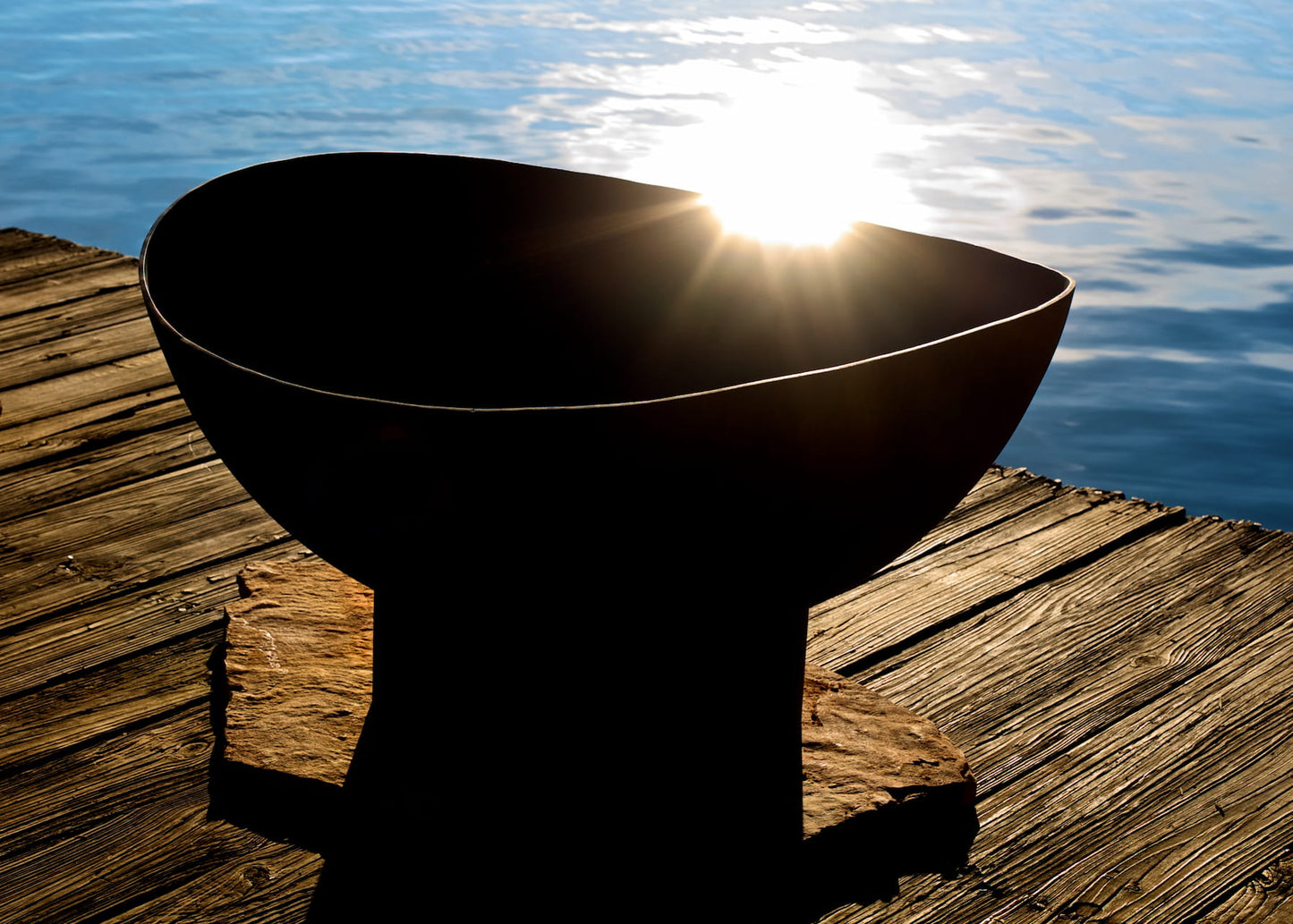 Scallop fire pit silhouetted by the setting sun on a dock with water in the background.