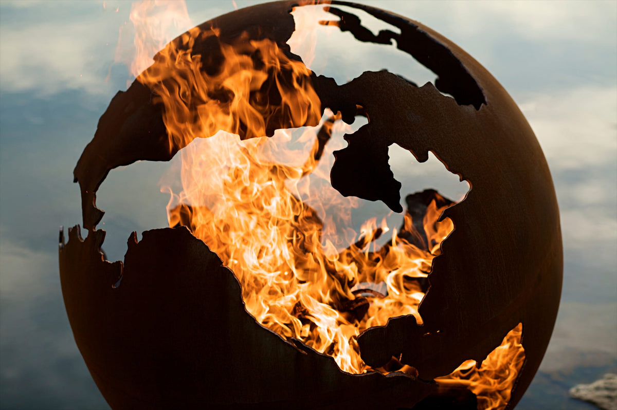 World map globe engulfed in flames against a cloudy sky. Made by Fire Pit Art in the USA.