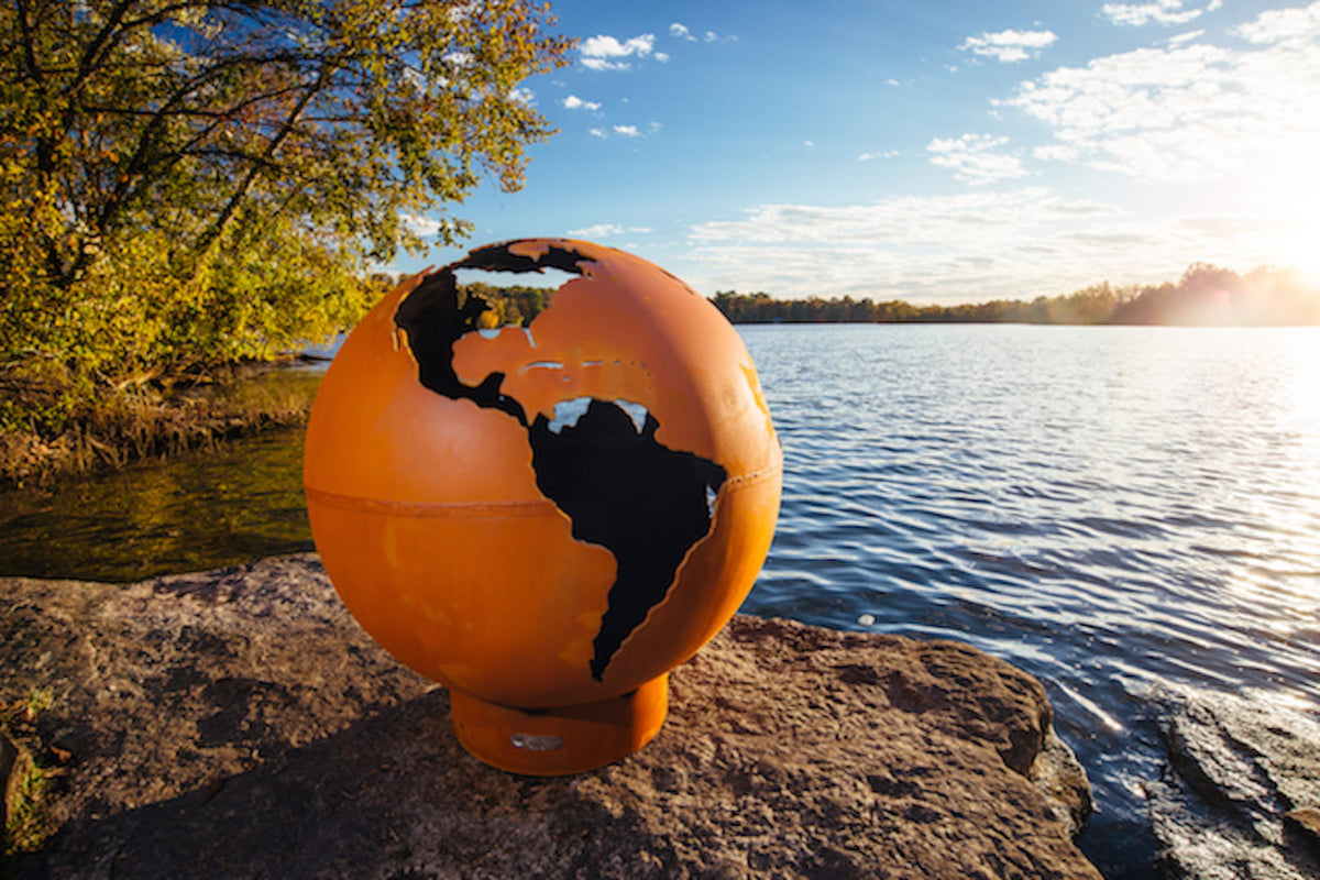View of the spherical Third Rock 36" carbon steel fire pit on a sunny day next to a lake.