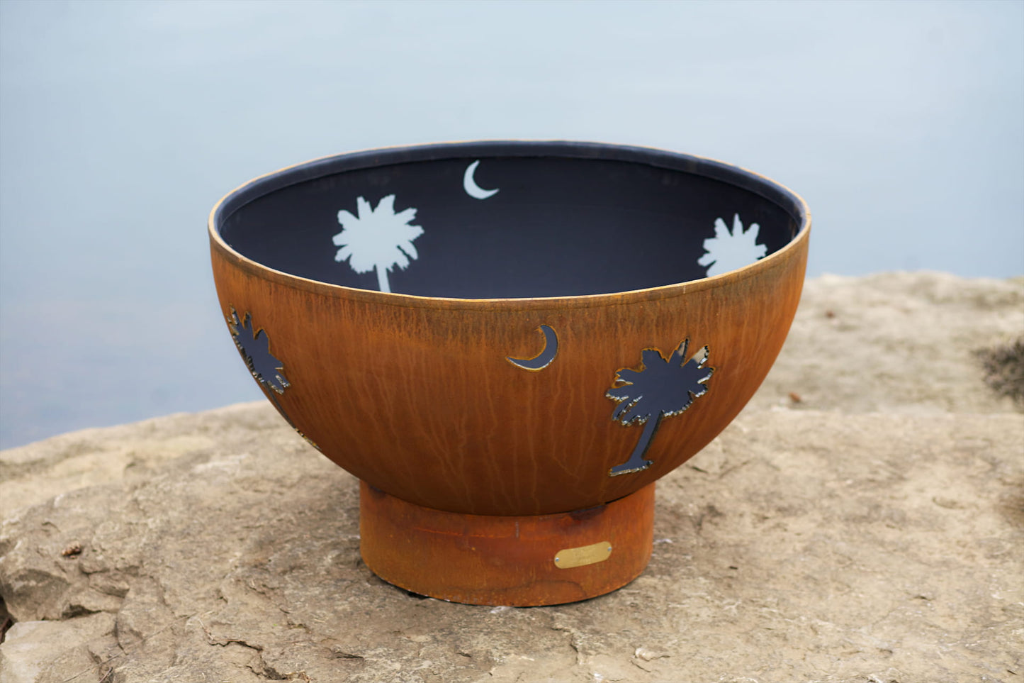 Tropical Moon 36-inch steel fire pit with palm tree and crescent moon cutouts, unlit, on a lakeside rock ledge.