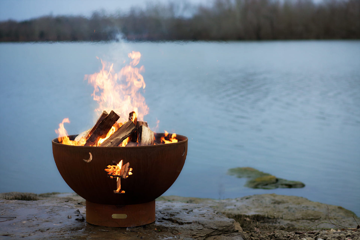 Fire Pit Art’s Tropical Moon 36-inch carbon steel fire pit with flames rising from burning logs; crescent and palm cutouts glowing by the lake. Made in USA.