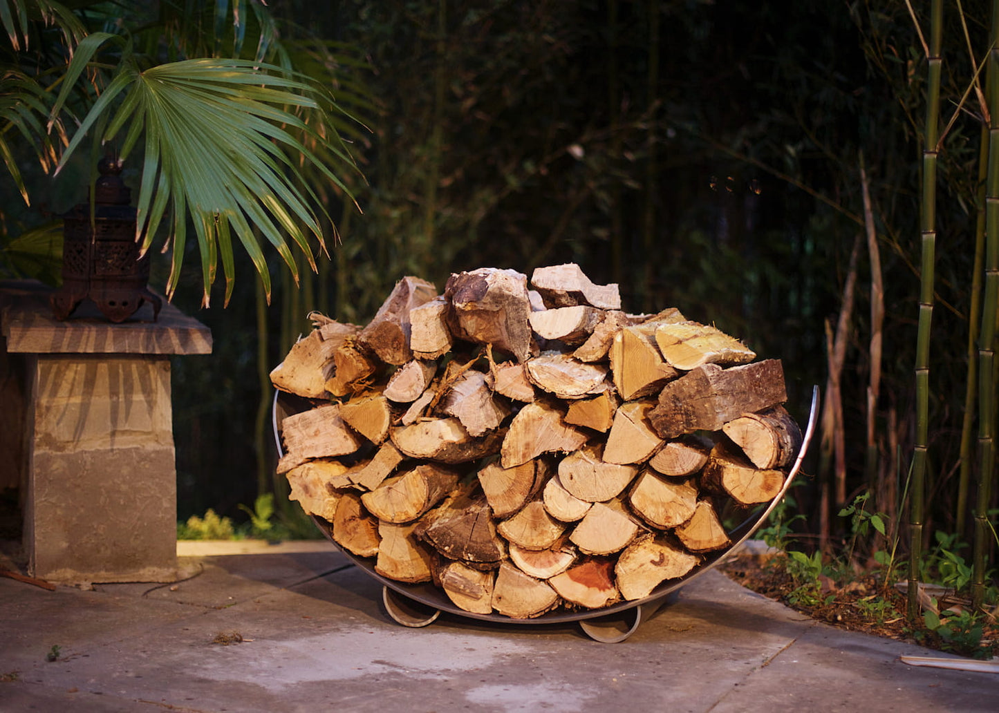Fire Pit Art’s stainless steel crescent-shaped log rack filled with wood on patio with greenery in background.