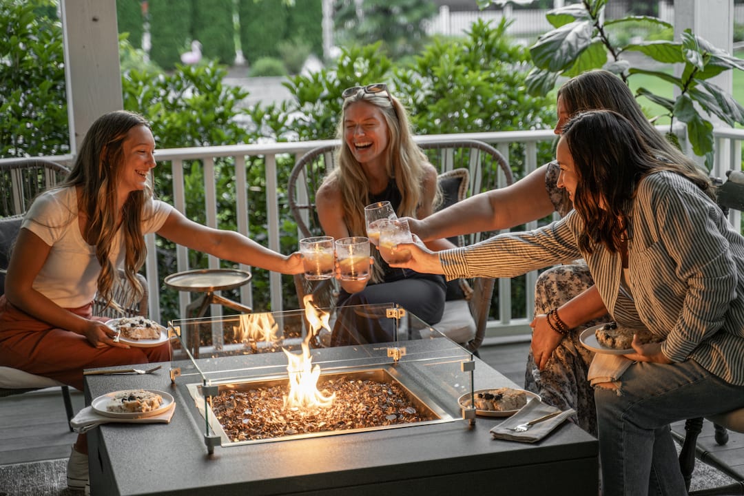 Four women laughing and toasting glasses around The Milan Square fire pit on a patio.