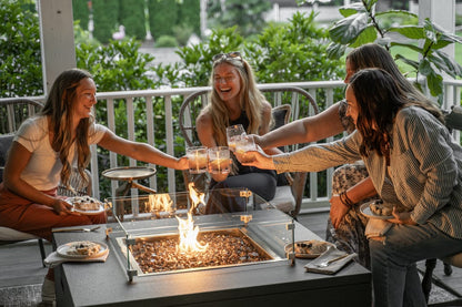 Four women laughing and toasting glasses around The Milan Square fire pit on a patio.