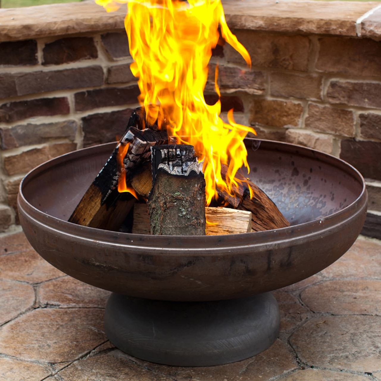 The Patriot fire pit with roaring flame against a brick wall backdrop on a patio.