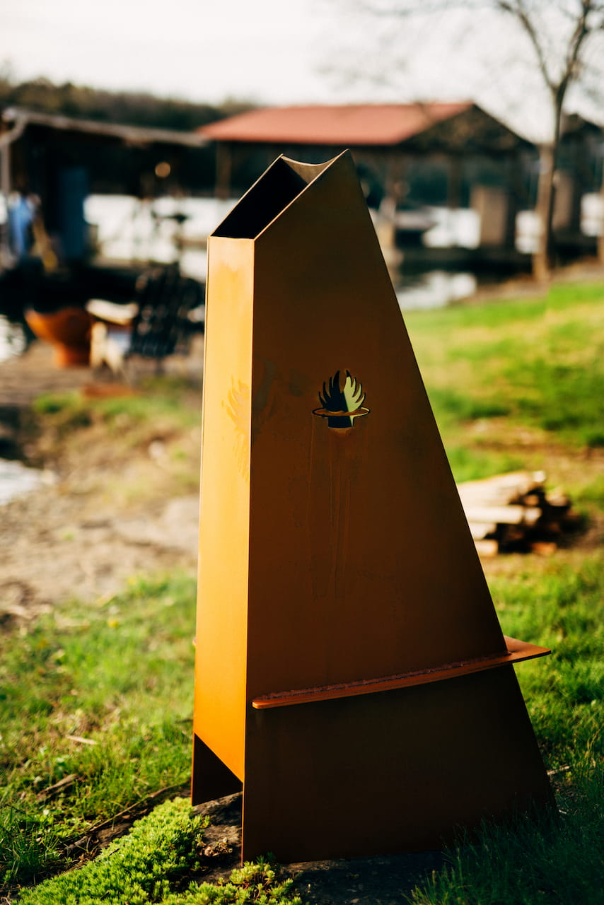 Back view of Vesuvius corten-steel outdoor chiminea with Fire Pit Art logo cutout and flue in a grassy yard.