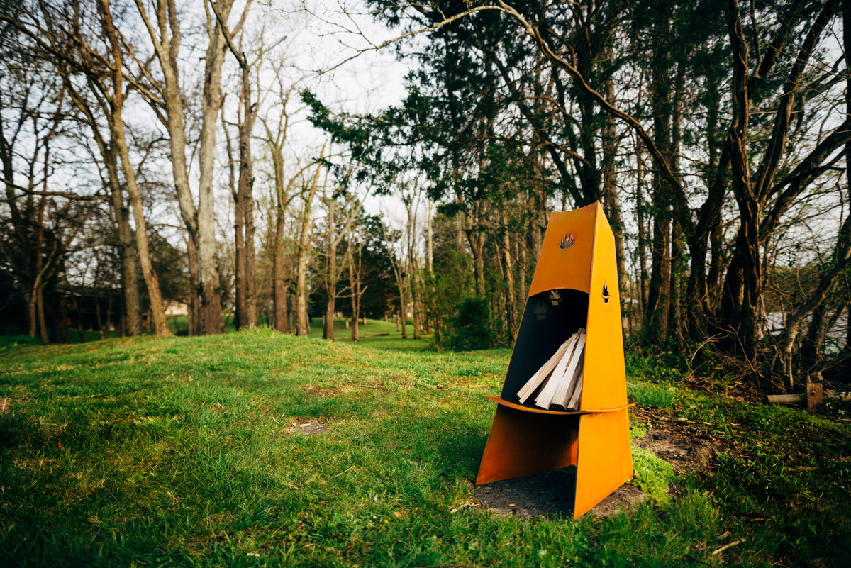 Wide lifestyle scene of the Vesuvius steel chiminea on a grassy lawn among trees, rich natural patina highlighting the sculptural shape.