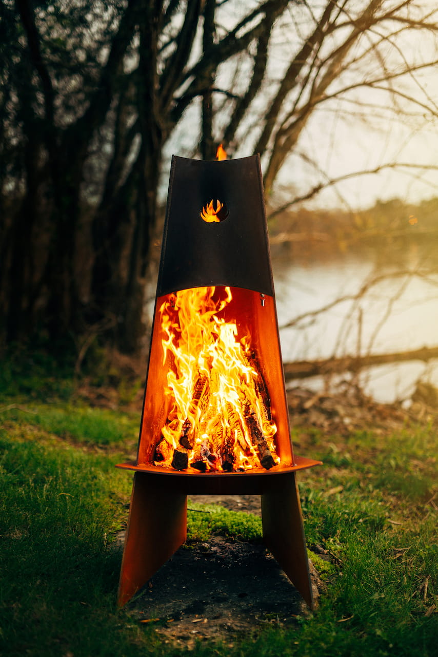 Fire pit with flames burning in a natural setting with trees and water in the background