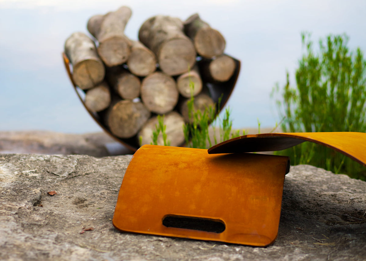 Sideview of disassembled two piece carbon steel Wings in Flight log rack showcasing the patina finish, and another one fully assembled with wood in it, in the background. By Fire Pit Art.