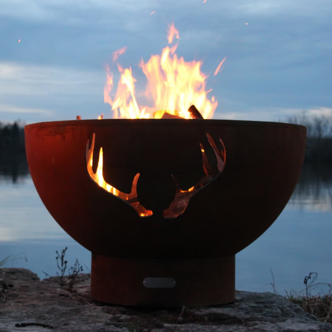 Antlers 36" Steel Fire Pit by Fire Pit Art at dusk with flames rising beside a lake. Available in wood burning, natural gas, or propane.