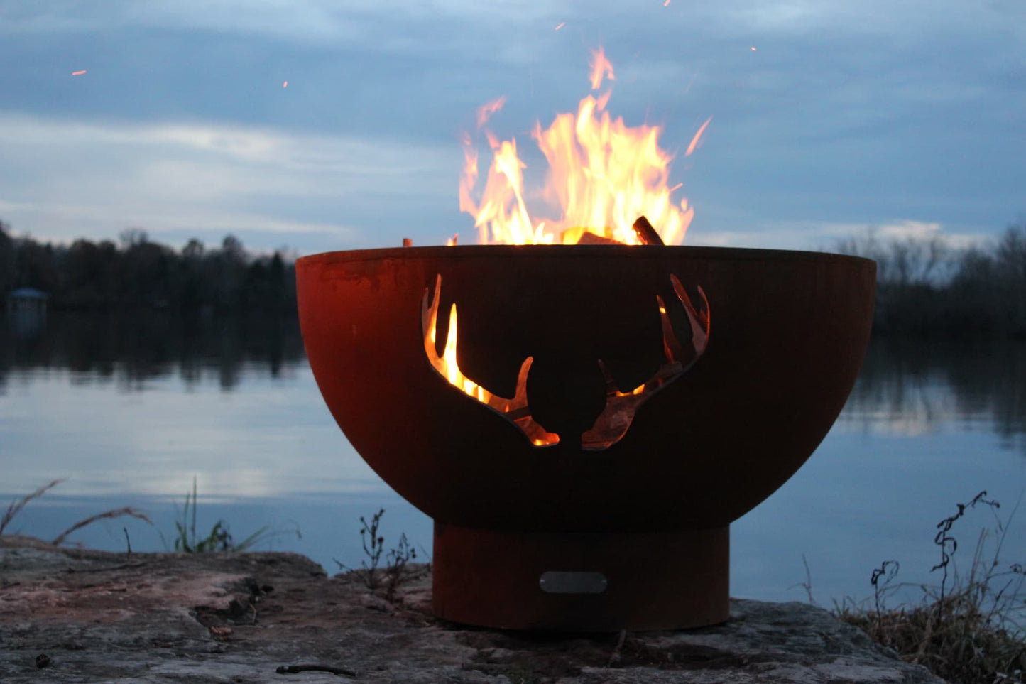 Wide view of Antlers 36" Steel Fire Pit with fire burning against a lakeside backdrop. Available in wood burning, natural gas, or propane.