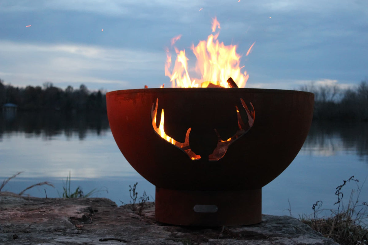Wide view of Antlers 36" Steel Fire Pit with fire burning against a lakeside backdrop. Available in wood burning, natural gas, or propane.