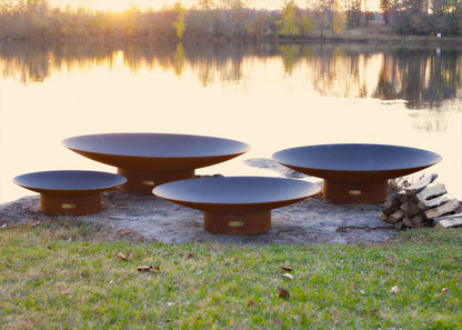 Side profile of all four sizes of the Asia Steel Fire Pit on rocky shore near dock.