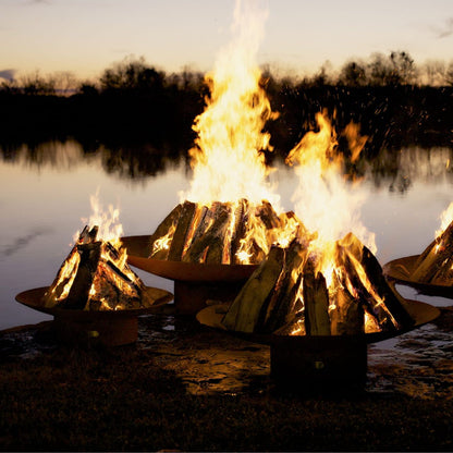 Three Asia Steel Fire Pits in varying sizes with wood burning at dusk.