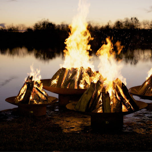 Three Asia Steel Fire Pits in varying sizes with wood burning at dusk.