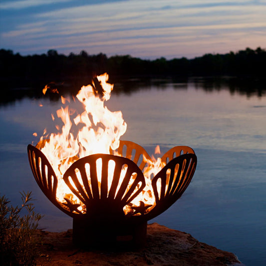 Barefoot Beach 42" Carbon Steel Fire Pit with seashell design glowing at sunset by the lake.  Made in the USA by Fire Pit Art.