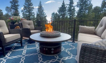 Outdoor patio with wine barrel fire pit + table ring surrounded by cozy chairs and blue patterned rug with trees in the background.