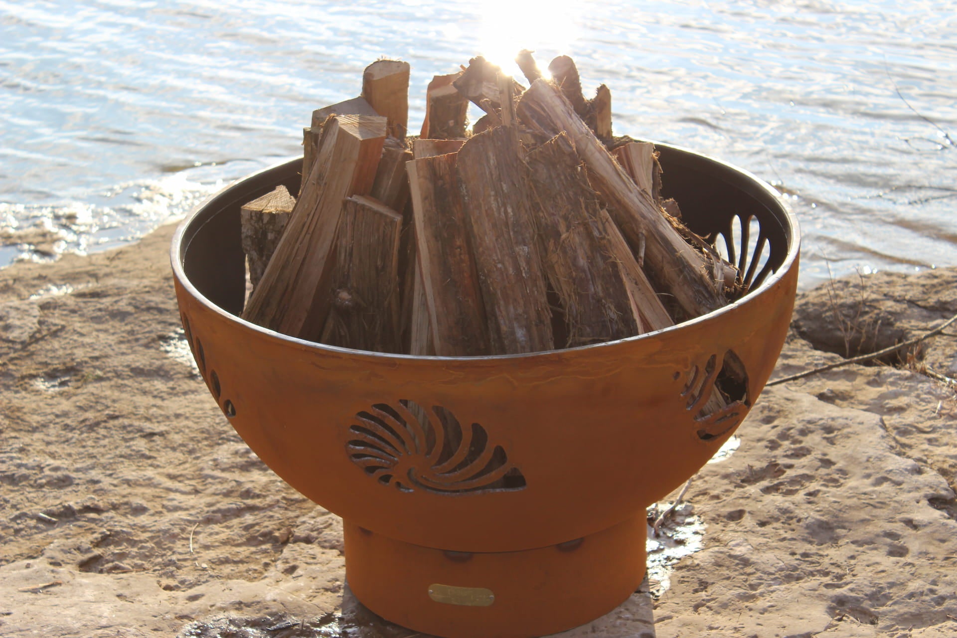 Beachcomber 36" carbon steel fire pit unlit and filled with wood, placed beside a lakeshore. By Fire Pit Art.