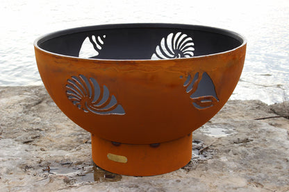 Full view of Fire Pit Art's Beachcomber 36" steel fire pit showing shell and conch designs in daylight.
