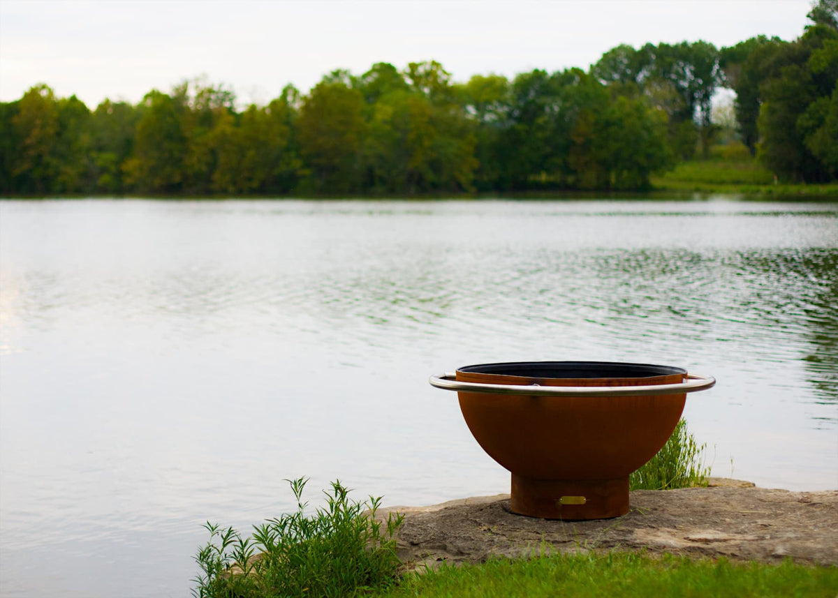 Bella Luna fire pit pictured from a distance by the lake’s edge. Made in USA by Fire Pit Art of carbon steel.