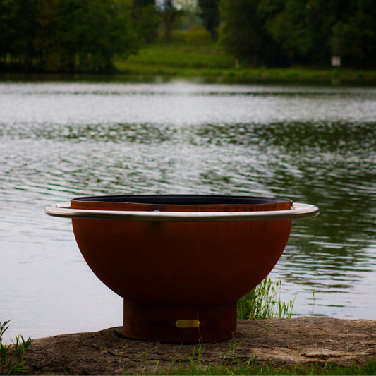 Bella Luna 48" carbon steel fire pit by Fire Pit Art on stone patio with lake and trees in background.  Made in USA by Fire Pit Art.