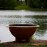 Bella Luna 48" carbon steel fire pit by Fire Pit Art on stone patio with lake and trees in background.  Made in USA by Fire Pit Art.