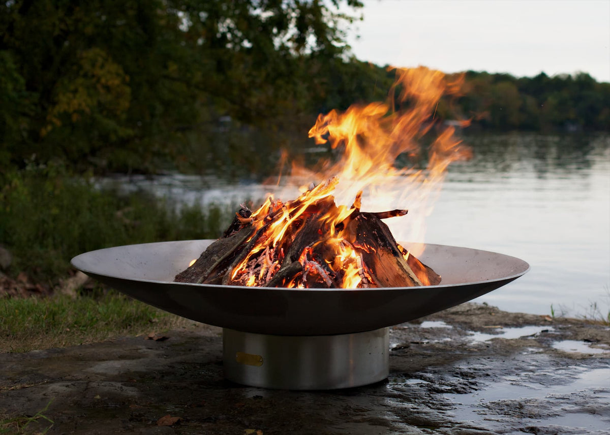 Bella Vita stainless steel fire pit burning brightly near wooded lake. Made in USA and comes in multiple sizes.