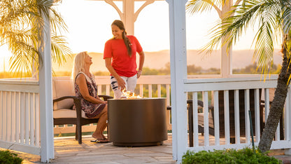 Two friends chat next to the Beverly Steel Powder Coated Fire Pit in a gazebo at sunset (pictured in Copper Vein).