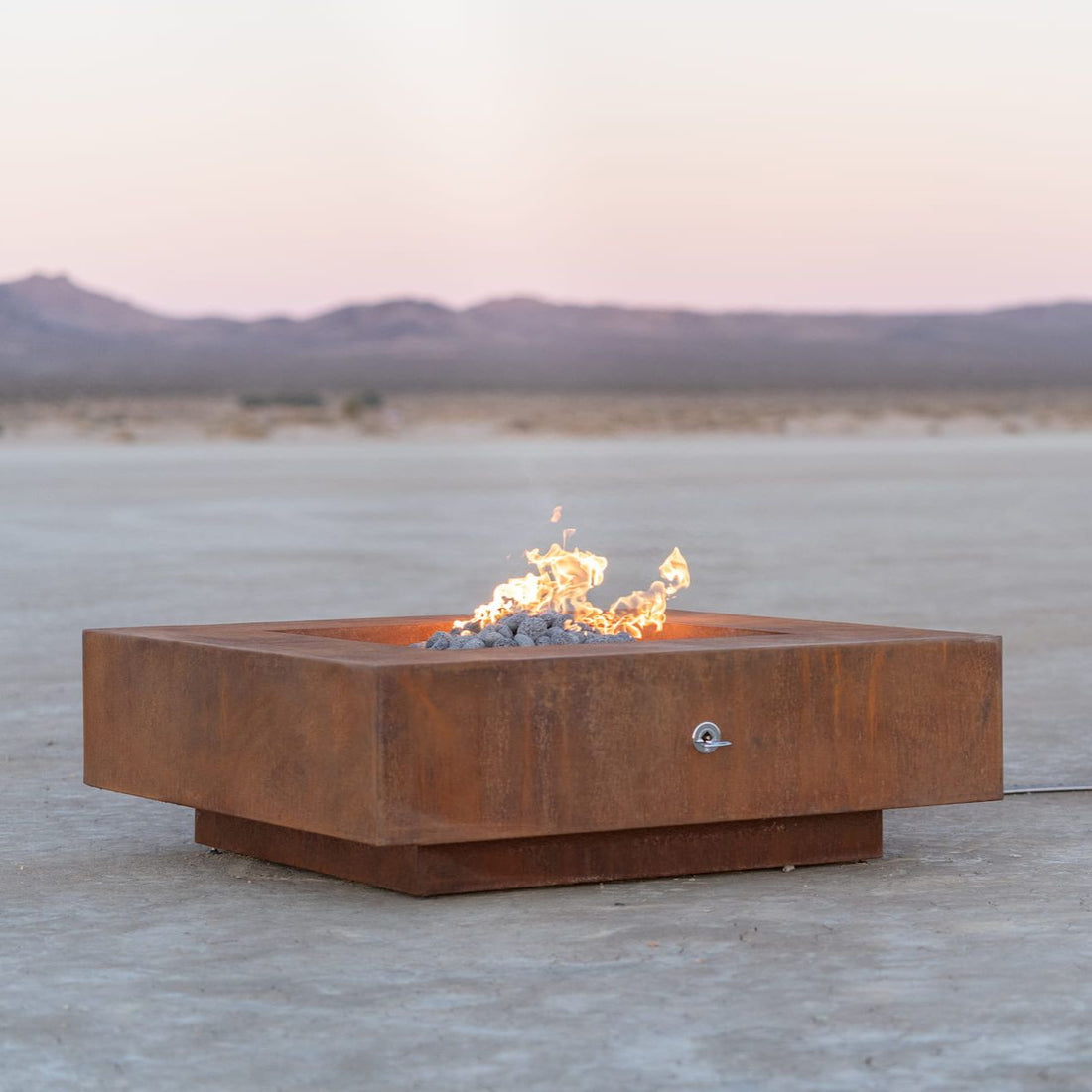Cabo square corten steel fire pit burning softly in a desert landscape at sunset, showing clean lines and modern design (shown in natural gas).