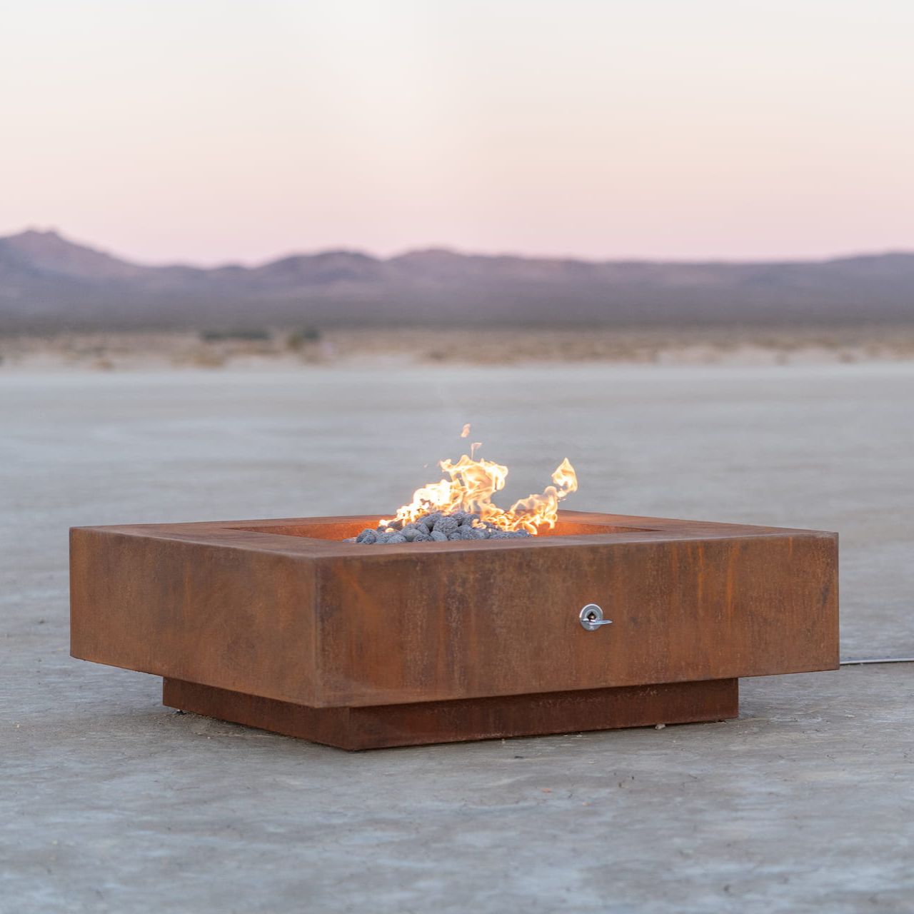 Cabo square corten steel fire pit burning softly in a desert landscape at sunset, showing clean lines and modern design (shown in natural gas).