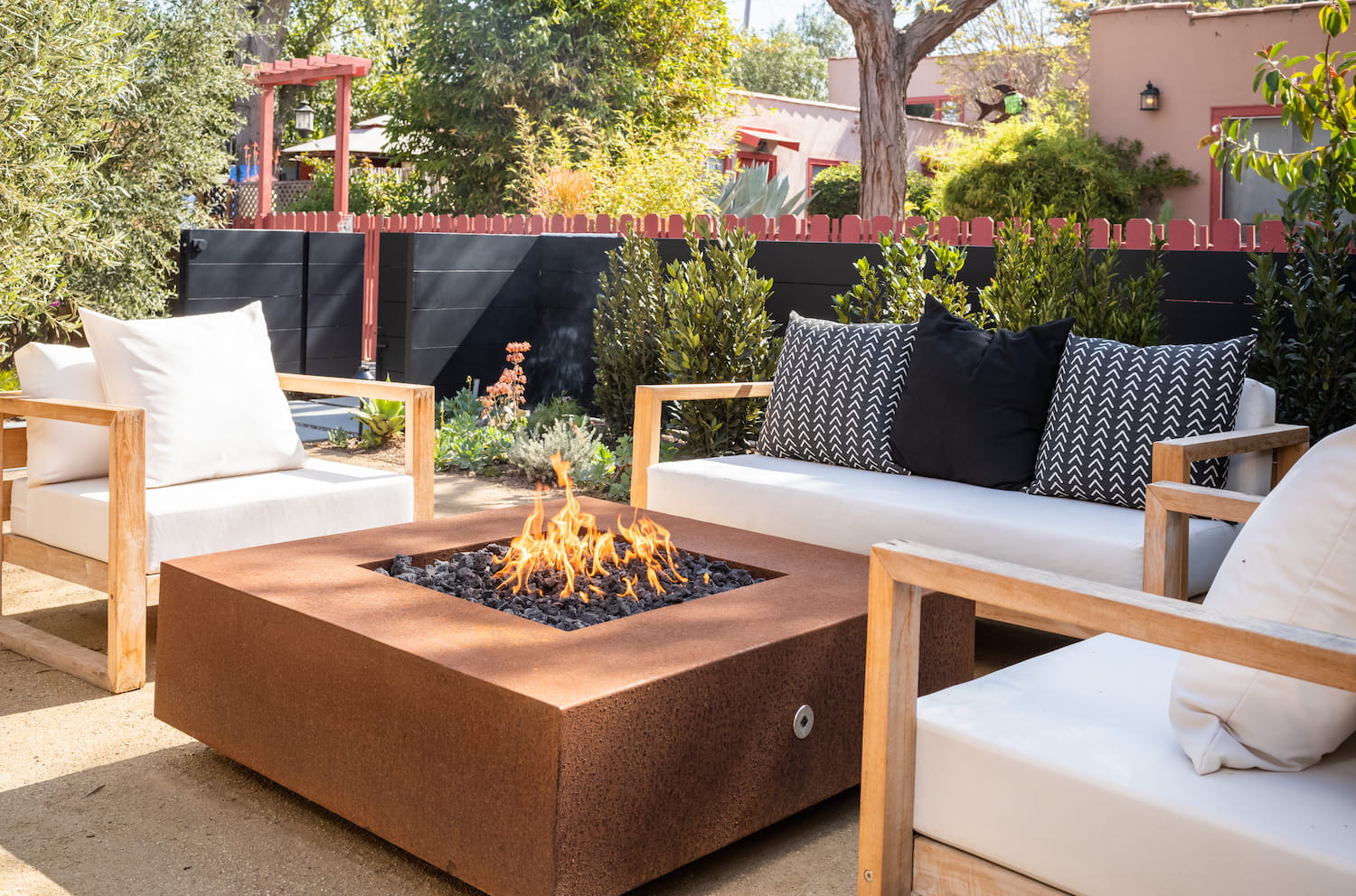Square Cabo fire pit in corten steel burning propane flames, centered between cushioned outdoor seating in a landscaped backyard.