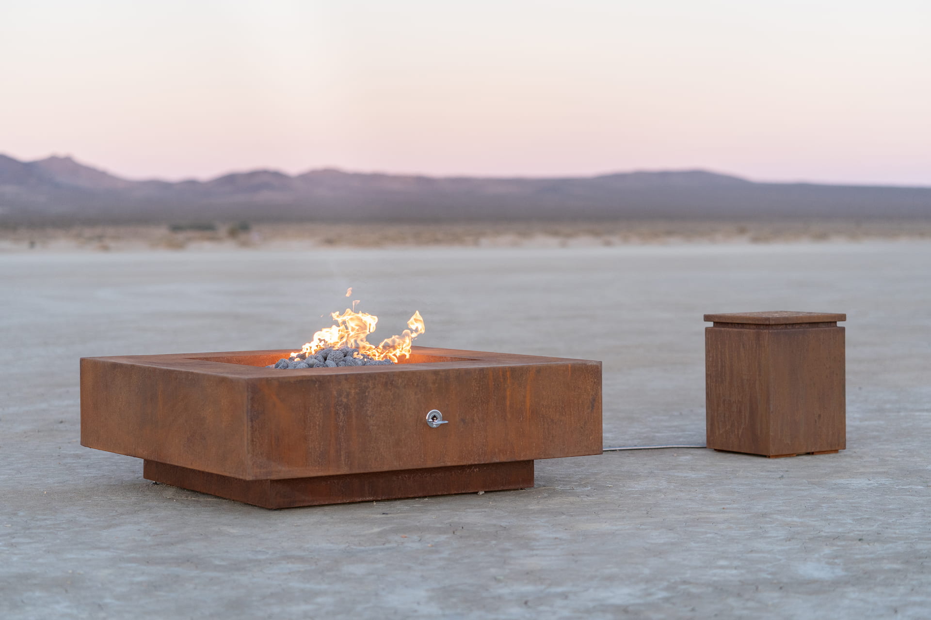 Cabo square corten steel fire pit with propane flame burning in an open desert landscape, paired with a matching propane tank enclosure.