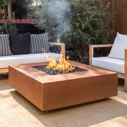 Cabo square corten steel fire pit with flames burning over lava rock on a modern outdoor patio with white seating (shown in natural gas).