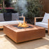 Square Cabo fire pit in corten steel burning propane flames, centered on a patio between cushioned outdoor seating and greenery.