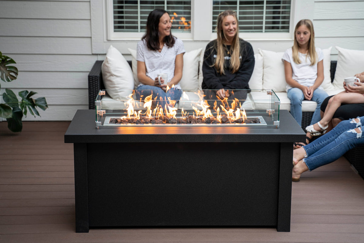 Front view of lit Carolina Evening Companion Propane Fire Table - Linear in Endura Graphite with family sitting around it (wind shield sold separately).

