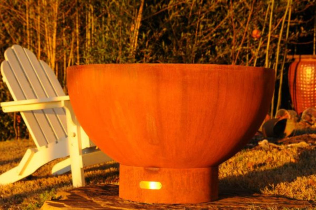 Side profile of Crater 36" carbon steel fire pit showing rounded bowl shape and pedestal base in golden hour light of garden setting.