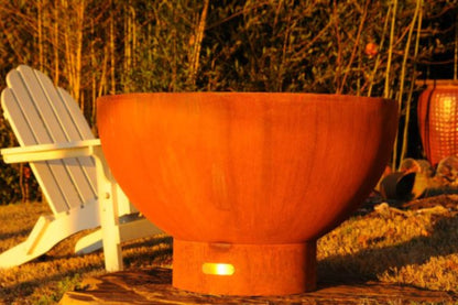 Side profile of Crater 36" carbon steel fire pit showing rounded bowl shape and pedestal base in golden hour light of garden setting.