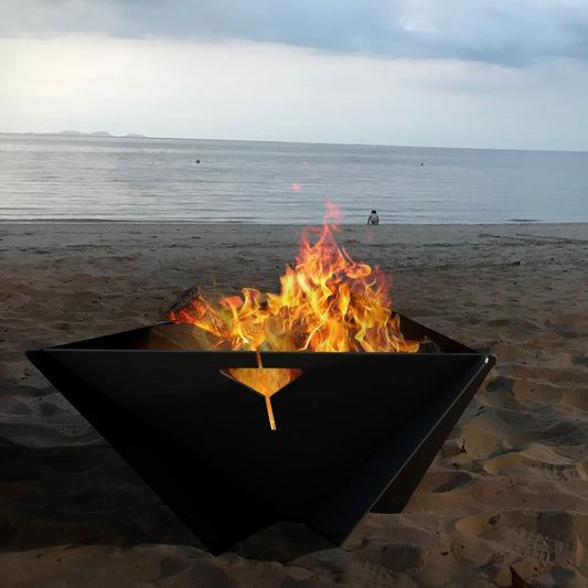 Up close image of steel Delta Fire Pit burning wood with the ocean at dusk in the background.