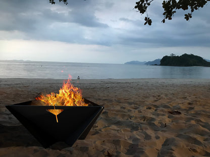 The Delta black steel fire pit burning wood on a beach with calm ocean water in the background.