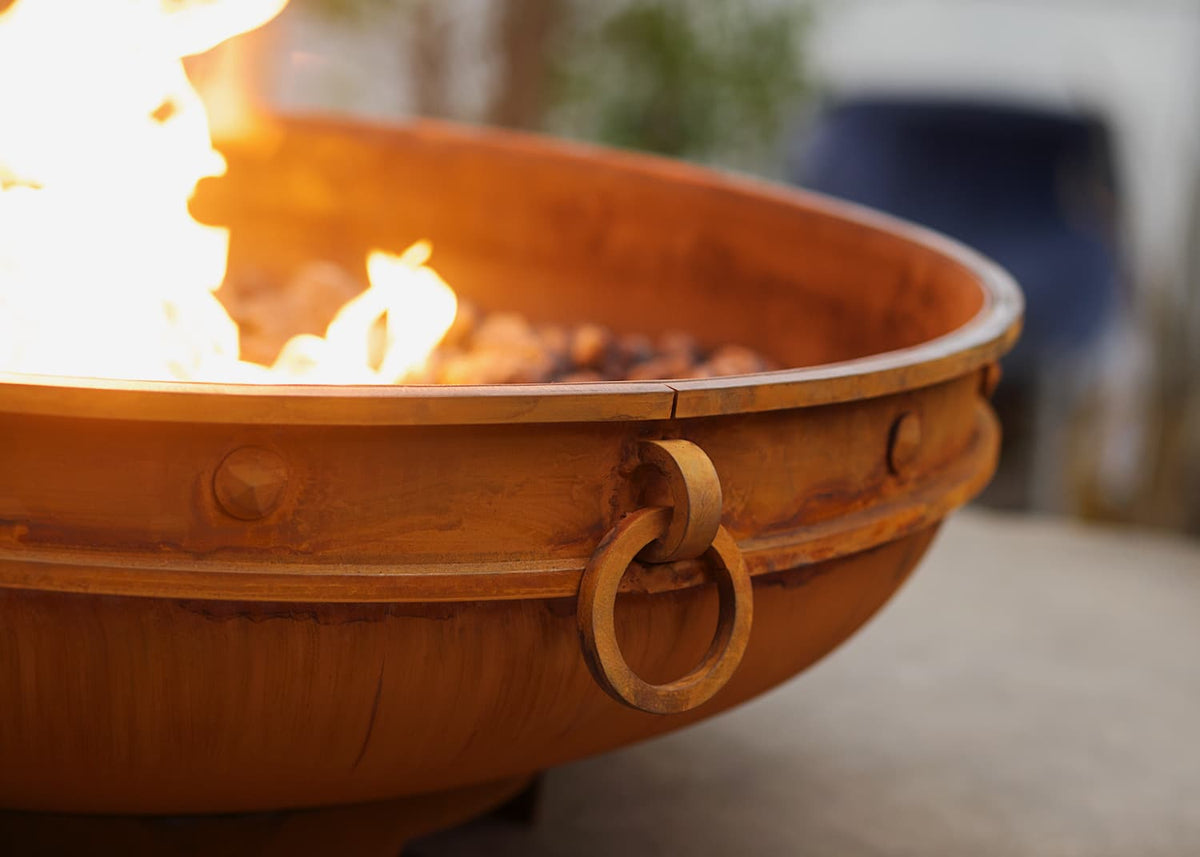 Close-up of Emperor 37" carbon steel fire pit with flames rising and detailed patina on steel surface with blurred background.