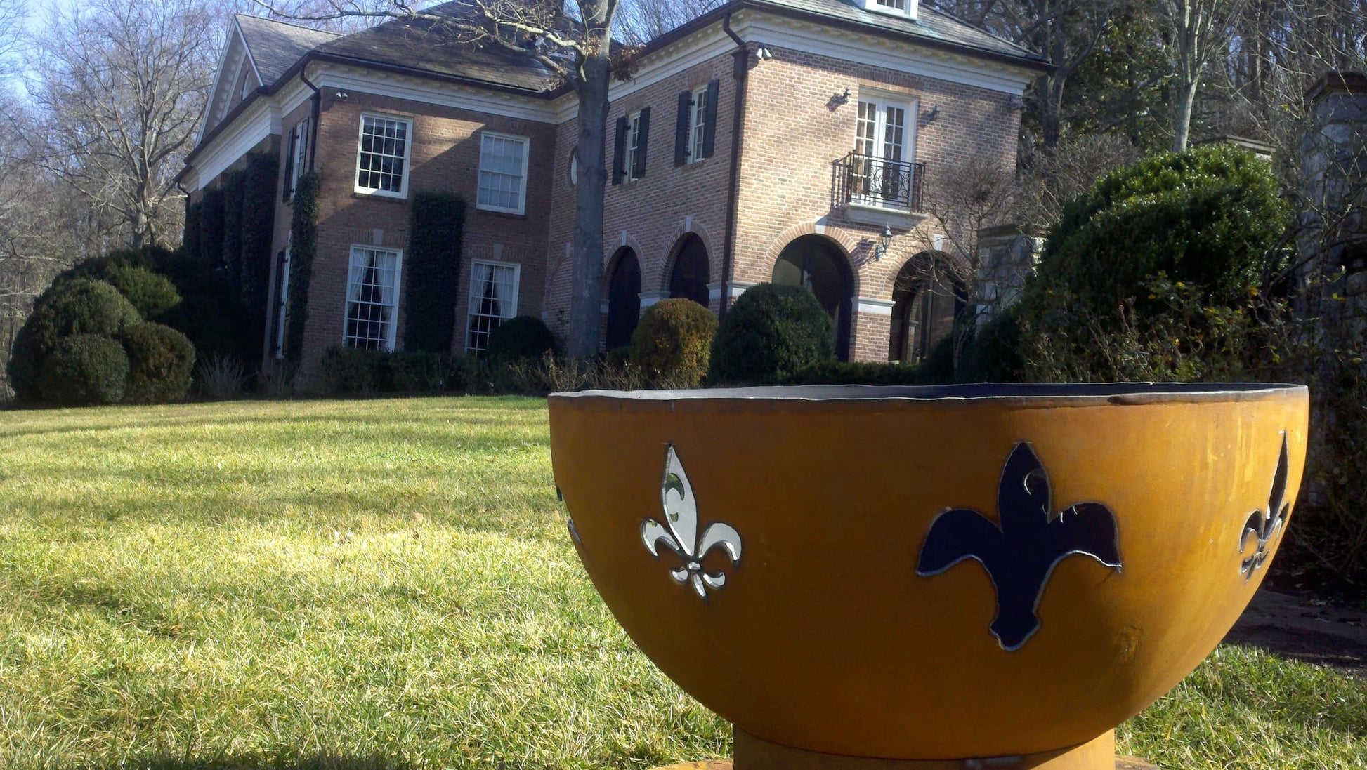 Side view of Fire Pit Art's fleur-de-lis 36" carbon steel fire pit in front of a traditional brick estate, showing patina finish and cutouts in daylight.