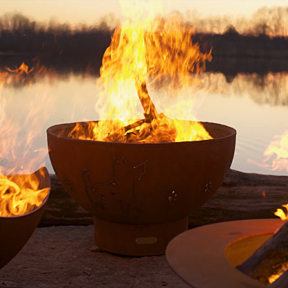 Funky Dog 36 " carbon steel fire pit by Fire Pit Art with dog and paw cutouts, lit at sunset on lakeside.