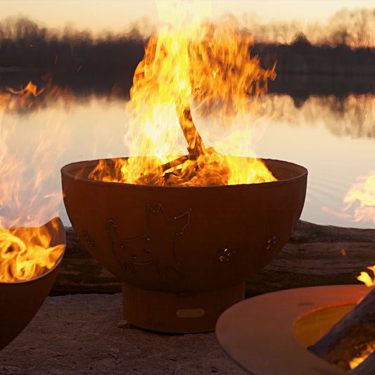 Funky Dog 36 " carbon steel fire pit by Fire Pit Art with dog and paw cutouts, lit at sunset on lakeside.