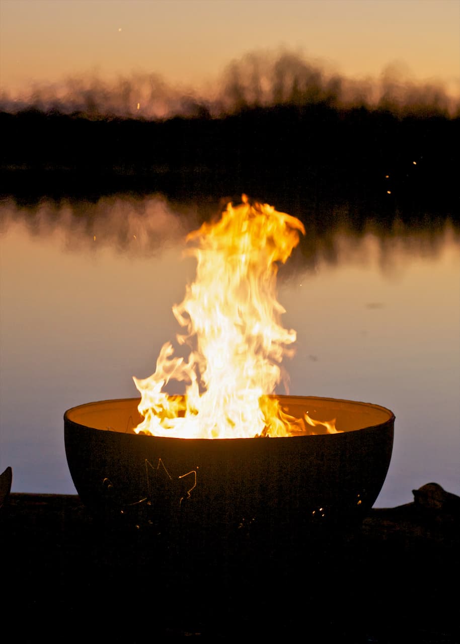 Funky Dog 36" carbon steel fire pit shown from side with tall flame and lake reflection. Made in the USA by Fire Pit Art.