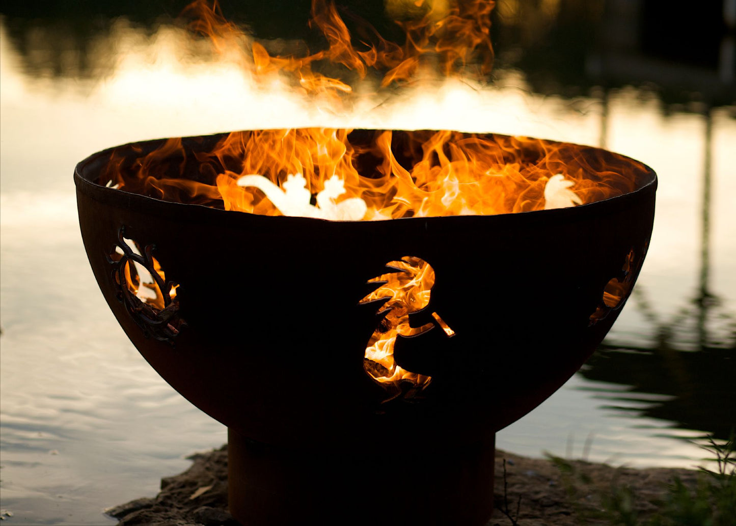 Fire burning through sun, kokopelli, and gecko cutouts on handcrafted steel fire pit by a lake at dusk.