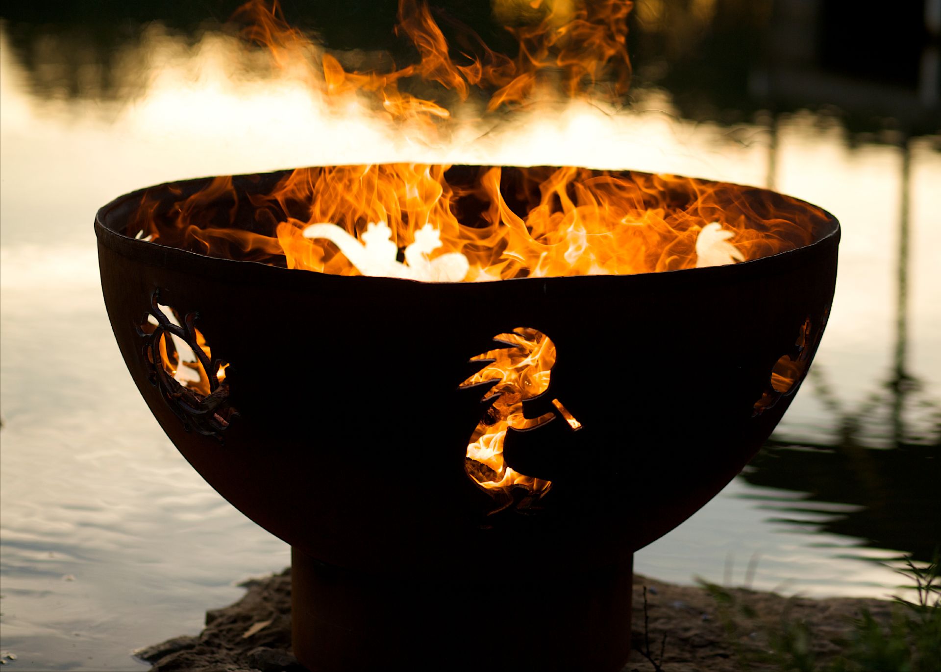Fire burning through sun, kokopelli, and gecko cutouts on handcrafted steel fire pit by a lake at dusk.